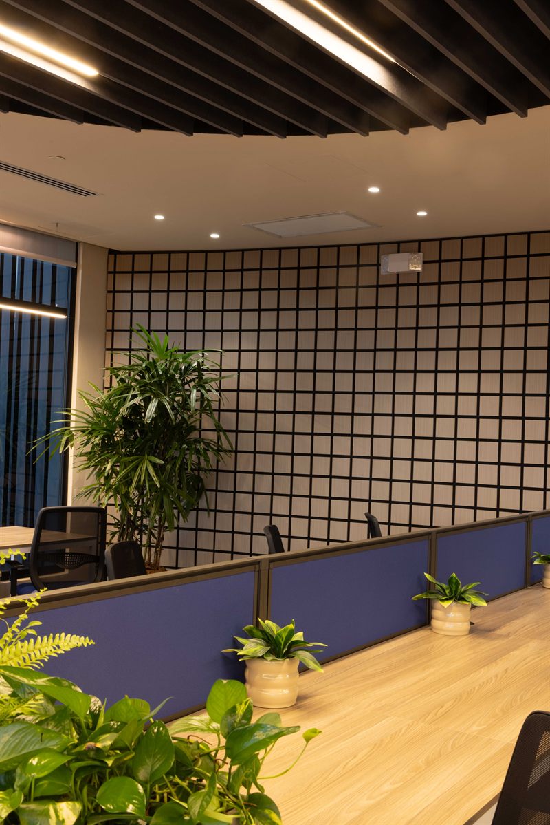 Área de trabajo bajo el cielo curvo de baffles, con luz lineal, muro en retícula y puestos separados por paneles azules