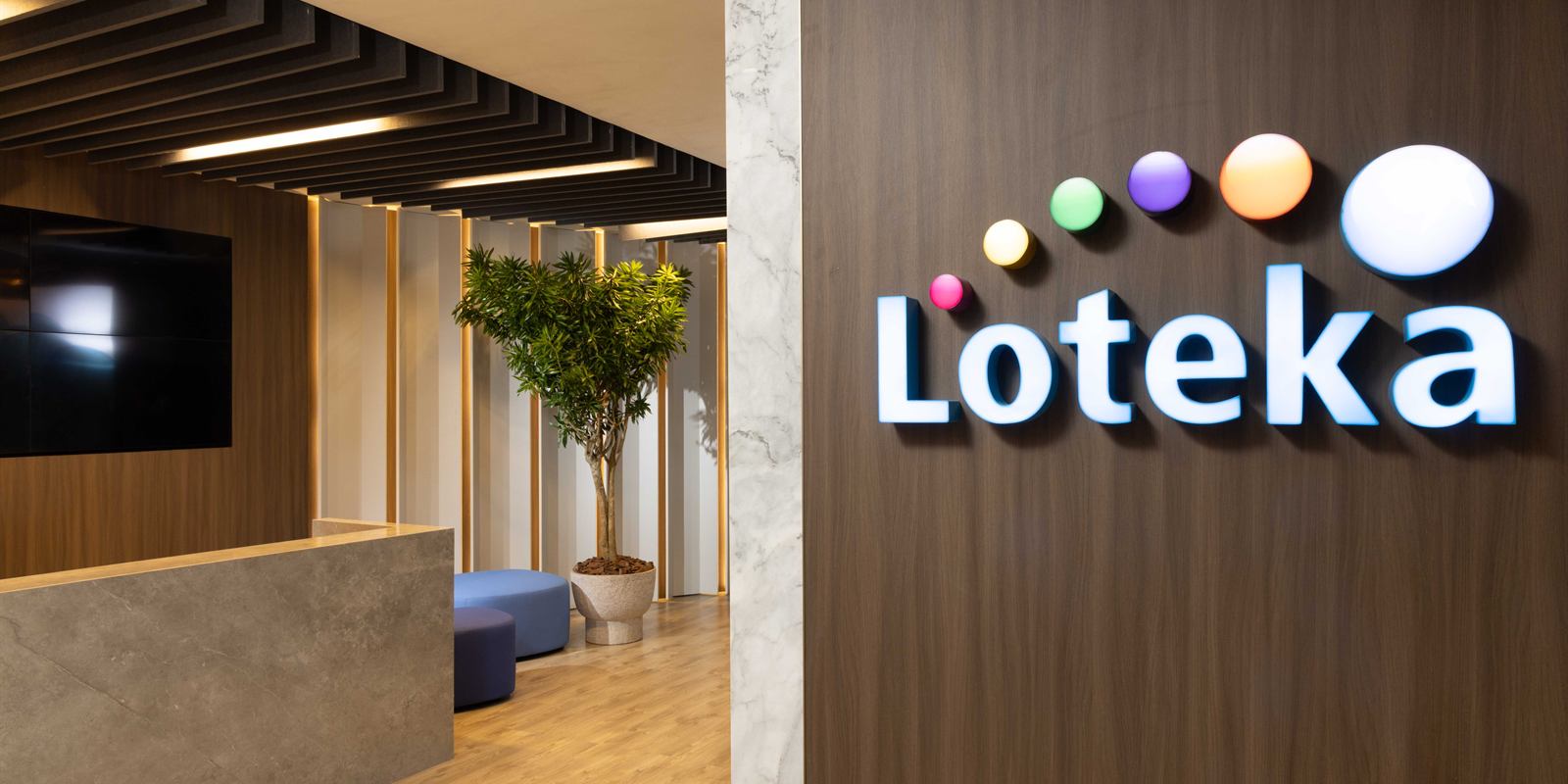Recepción de la oficina corporativa: letrero iluminado de Loteka sobre un muro de paneles con acabado de madera, mostrador y cielo de baffles de fieltro
