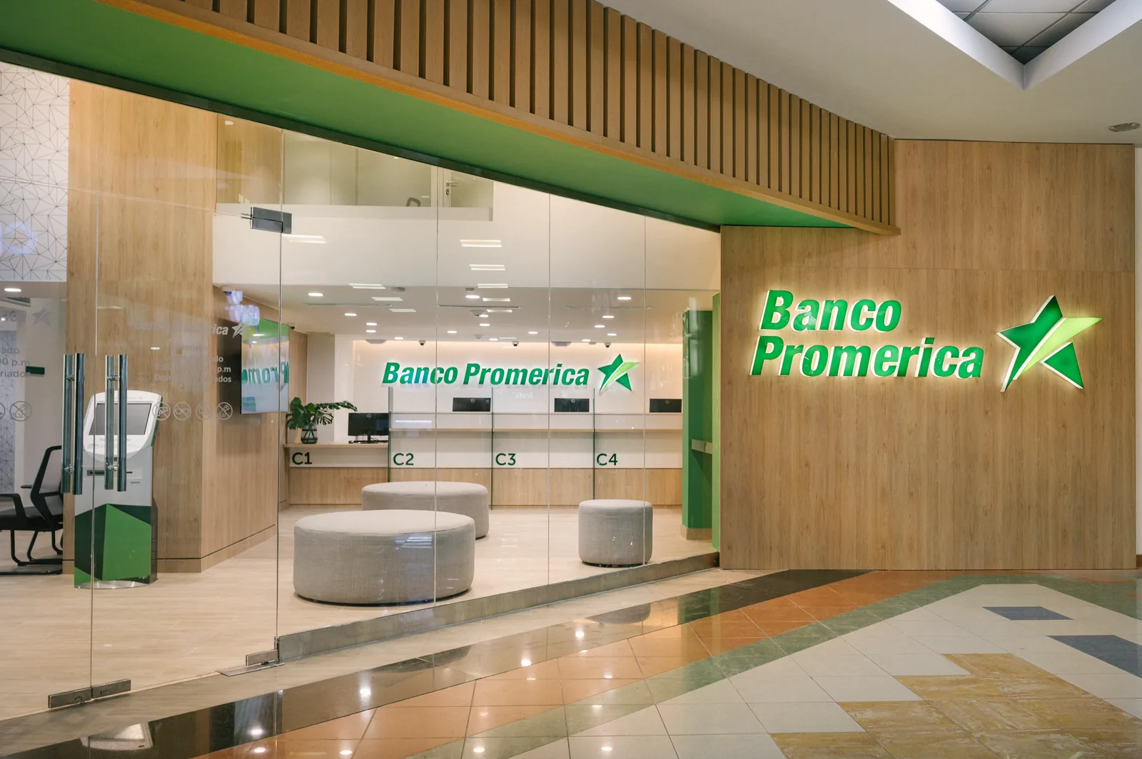 Frente de la sucursal hacia la plaza interior: muro cortina de vidrio, rótulo del banco sobre un plano de madera y, encima del vano, una franja de listones de madera