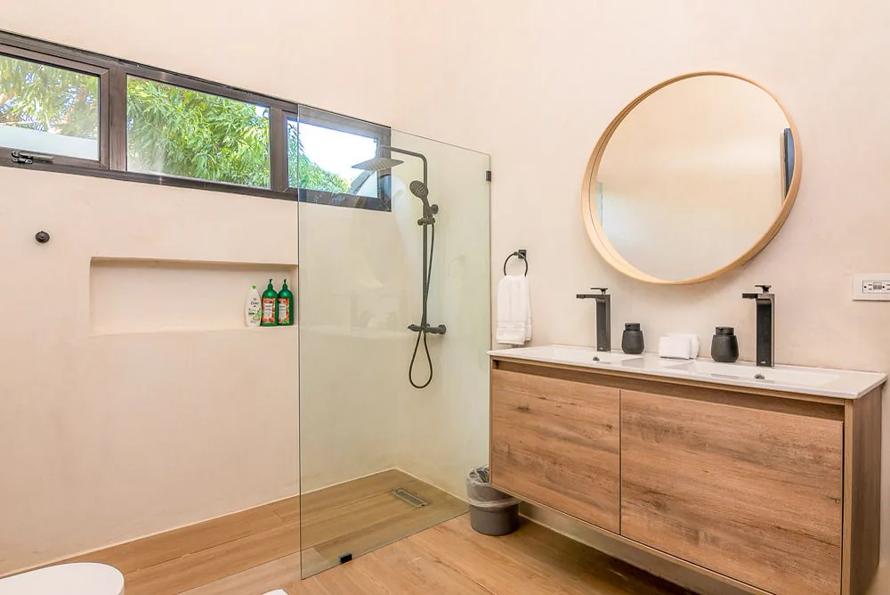 Baño con mueble suspendido de madera, doble lavamanos, espejo redondo y ducha corrida con ventana alargada