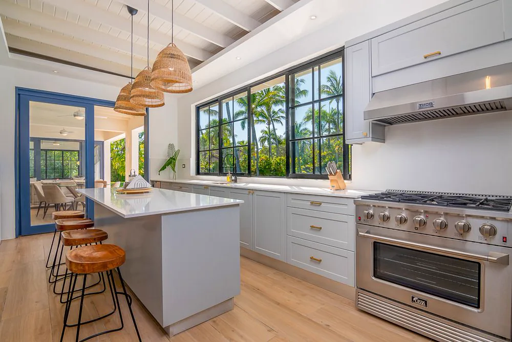 Cocina con isla central de tope claro, taburetes, lámparas de fibra y ventanas de carpintería azul hacia el jardín