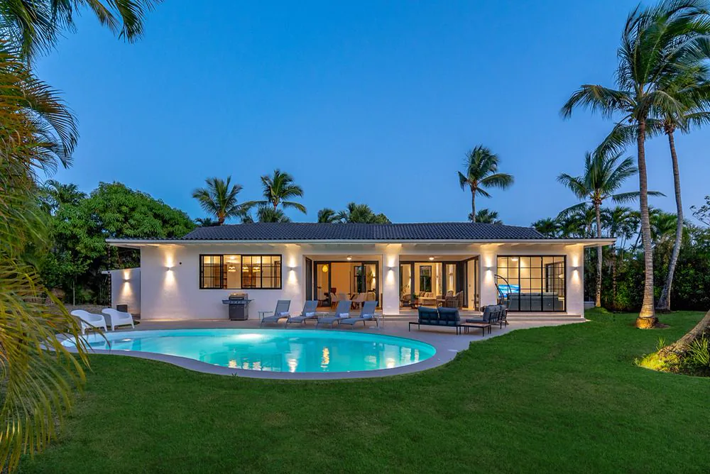 La villa al atardecer, con las luces encendidas detrás de los ventanales, la piscina en primer plano sobre el césped y las palmas contra el cielo azul