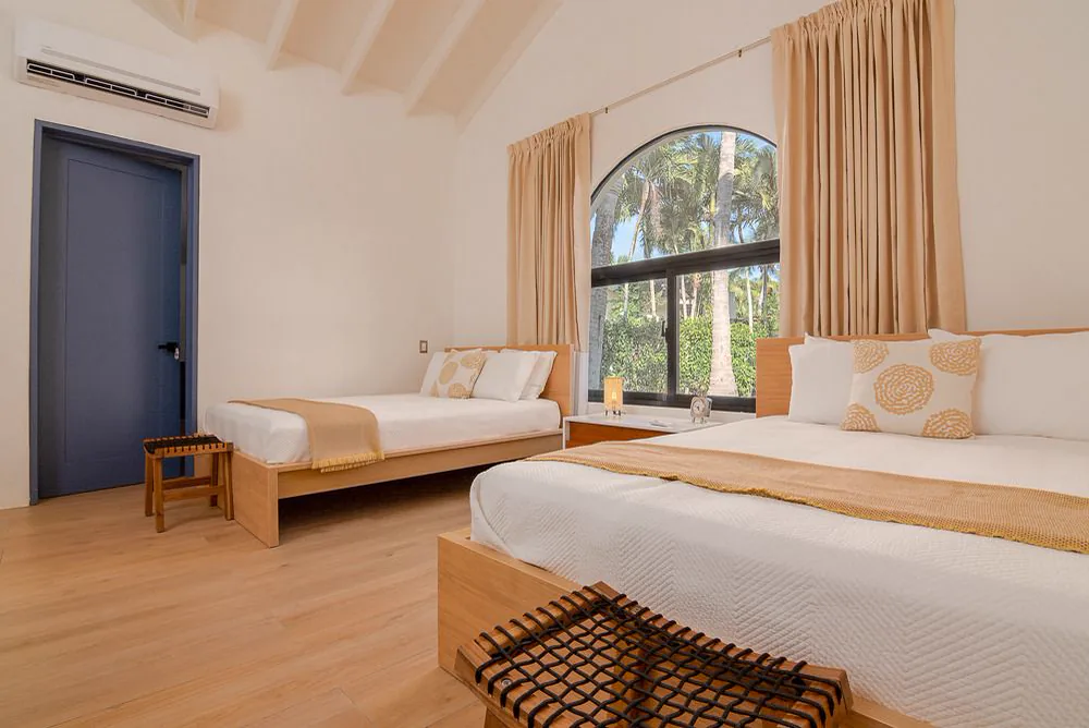 Habitación con dos camas bajo la cubierta inclinada, con una ventana en arco hacia las palmas y cortinas claras