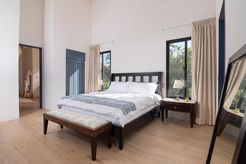 Habitación principal de techo alto, con cama de madera oscura, banqueta a los pies y una ventana alta hacia el jardín
