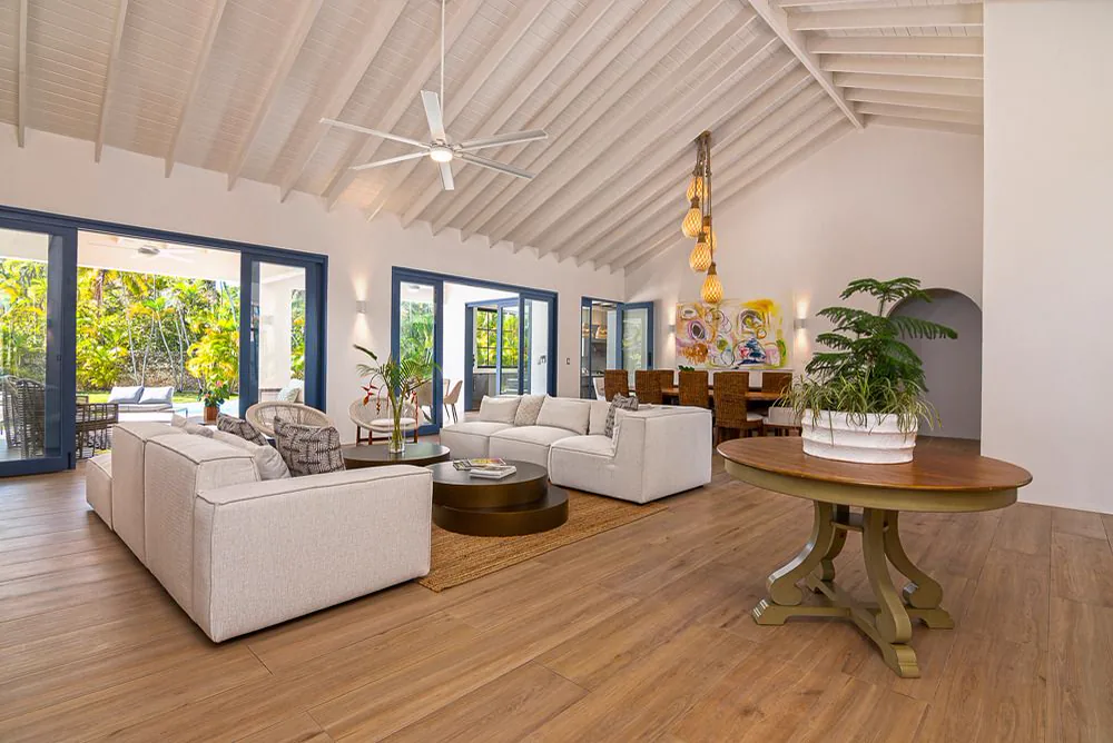Sala principal bajo la cubierta inclinada de madera pintada de blanco, con ventilador de techo, mesa redonda de madera y ventanales hacia la vegetación