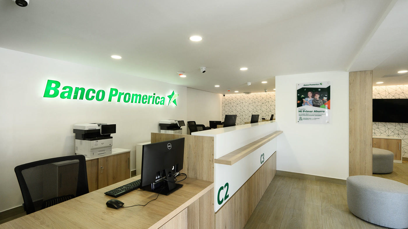 Sal&oacute;n terminado de la sucursal de Banco Promerica en Gustavo Mej&iacute;a Ricart: el r&oacute;tulo iluminado sobre el muro blanco, los puestos de caja C2 y C3 y el &aacute;rea de espera al fondo